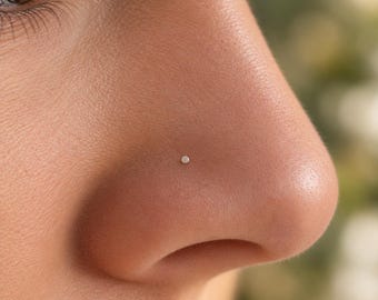 May include: A small, circular, silver-toned nose stud featuring a flat, polished surface. The jewelry is positioned centrally on the side of the nose, presenting a minimalist and understated appearance with a smooth, reflective metallic finish.