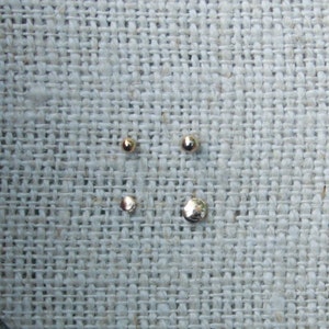 May include: Four small, shiny, gold-colored metal spheres of varying sizes are arranged on a textured, light-colored fabric. The spheres appear to be jewelry components, possibly beads or studs.