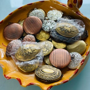 May include: A collection of assorted metal lockets in a yellow ceramic bowl with red spots. The lockets feature various finishes including copper, gold, and silver tones, with textured, floral, and geometric patterns. Some are in plastic bags.
