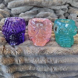 May include: Three decorative skull-shaped containers in shades of purple, pink, and teal. The containers are made of a translucent material, allowing light to pass through. The skulls are resting on a gray, textured surface.