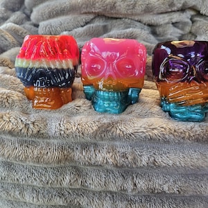 May include: Three colorful skull-shaped decorative objects. The skulls are painted with vibrant gradients of red, orange, blue, and purple. The objects are sitting on a soft, gray, textured surface.