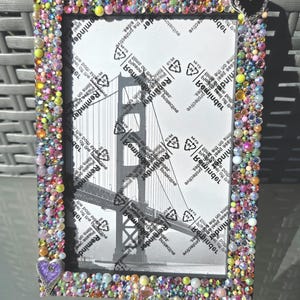 May include: A rectangular picture frame featuring a border densely covered in multicolored, iridescent beads and small rhinestones. A silver-toned heart charm is attached to the top right and bottom left corners. The frame contains a protective film.