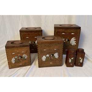 May include: A set of six wooden boxes with a rooster design. The boxes are various sizes, with a dark brown finish and hand-painted rooster illustrations in white, black, and gold. The set includes two small spice jars.