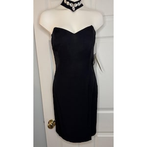 May include: A black strapless dress with a sweetheart neckline. The dress is displayed on a mannequin. A black choker necklace with large, clear rhinestones is around the mannequin's neck. The dress has a knee-length hemline.