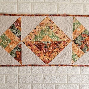Op de afbeelding: Een rechthoekige gewatteerde tafelloper met een reeks geometrische driehoekige en ruitvormige patches. De stof toont een mix van herfstachtige bloemen- en bladpatronen in tinten oranje, groen en bruin, geplaatst tegen een crèmekleurige achtergrond.