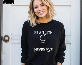 Pode incluir: Uma camisola preta de manga comprida com o texto branco BE A LILITH NEVER EVE, centrado sobre um gráfico branco de uma lua crescente combinada com um símbolo de adaga. A peça tem gola redonda e mangas compridas.