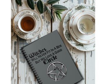 May include: A gray spiral-bound notebook with the text "Witches Will Never Fit In A Box But We Love A Good Circle" and a pentagram design. The notebook is on a distressed white wooden surface, with teacups and plates.