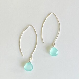 Pode incluir: Um par de brincos prateados com uma pedra preciosa azul clara em forma de gota. Os brincos t&ecirc;m um design simples e curvo, com a pedra preciosa pendurada na parte inferior. O fundo &eacute; branco.