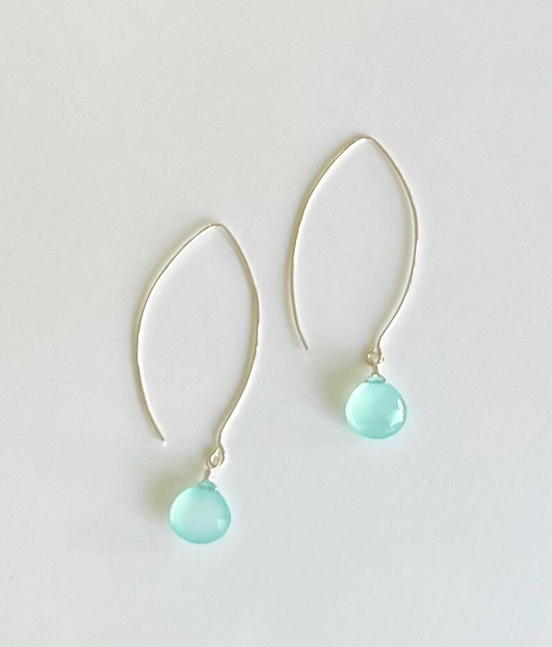 Pode incluir: Um par de brincos prateados com uma pedra preciosa azul clara em forma de gota. Os brincos t&ecirc;m um design simples e curvo, com a pedra preciosa pendurada na parte inferior. O fundo &eacute; branco.