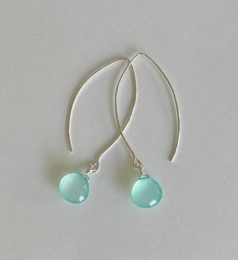Pode incluir: Um par de brincos pendurados elegantes com pedras preciosas em forma de gota, azul claro, suspensas por fios de prata curvos. Os brincos s&atilde;o exibidos em um fundo branco liso, destacando seu design delicado e cor.