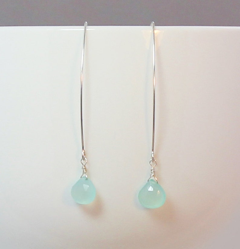 Pode incluir: Um par de brincos pendurados elegantes. Cada brinco apresenta um fio longo e fino de prata com uma pequena pedra preciosa em forma de l&aacute;grima azul clara na parte inferior. As pedras preciosas t&ecirc;m uma superf&iacute;cie facetada, que capta a luz.