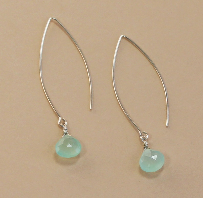 Pode incluir: Um par de brincos pendurados prateados. Cada brinco apresenta um design de fio curvo e alongado com uma pequena pedra preciosa verde-claro facetada em forma de gota pendurada na parte inferior. As pedras preciosas s&atilde;o de uma cor verde pastel suave.