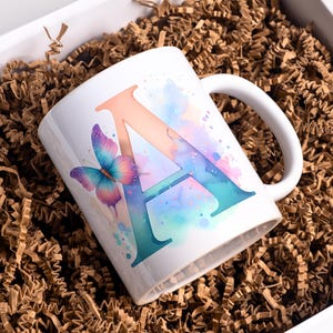 May include: White ceramic mug with a large letter "A" in coral and teal watercolor design, and a colourful butterfly. The mug is presented in a box with brown crinkle paper. A great gift.
