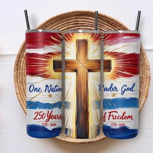 May include: Three stainless steel tumblers with black straws, featuring a patriotic design. The design includes a wooden cross, red and blue stripes, and the text "One Nation Under God" and "250 Years of Freedom 1776-2026". The tumblers are displayed on a woven tray.