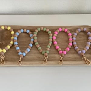 Könnte beinhalten: Fünf Schlüsselanhänger mit Perlen in Gelb, Blau, Grün, Pink und Lila. Jeder Schlüsselanhänger hat Holzperlen und einen goldfarbenen Verschluss. Die Schlüsselanhänger werden auf einem Holztablett präsentiert.