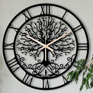 Peut inclure: Une horloge murale en métal noir avec un motif d'arbre de vie. L'horloge comporte des chiffres romains et des aiguilles dorées. L'horloge est fixée sur un mur gris clair. Le diamètre de l'horloge est d'environ 60 cm.
