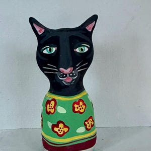 May include: A whimsical black cat figurine with green eyes and a pink nose. The cat wears a green shirt adorned with red and yellow floral patterns. The base of the figurine features horizontal stripes of pink, red, and yellow.