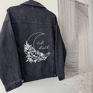 May include: Black denim jacket with a white floral and crescent moon design on the back. The text "Til death 10.24.20" is printed within the design. The jacket is hanging on a black hook.