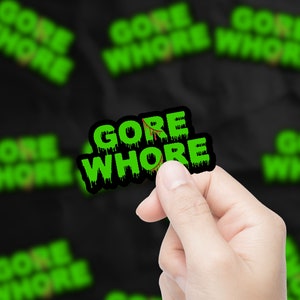 May include: A green and black sticker with the text "GORE WHORE" in dripping green letters. The sticker is being held by a hand.
