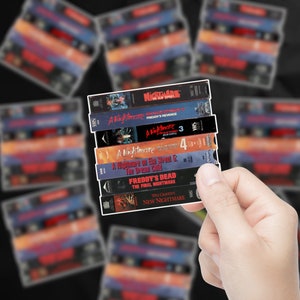 May include: A stack of VHS tapes featuring the horror movie franchise "A Nightmare on Elm Street". The tapes are arranged vertically, with the titles "A Nightmare Freddy's Revenge", "A Nightmare 3", "A Nightmare 4", "A Nightmare on Elm Street 5: The Dream Child", "Freddy's Dead: The Final Nightmare", and "Wes Craven's New Nightmare" visible.
