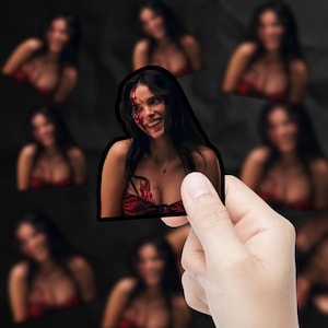 May include: A sticker featuring a smiling woman with long dark hair, blood splatters on her face and chest, and wearing a red top. The sticker is held by a hand, with blurred images of the same woman in the background.