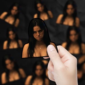 May include: A sticker featuring a woman with long dark hair and a pendant necklace, with a slightly sad expression. The sticker is held in a hand, with other blurred images of the same woman in the background.