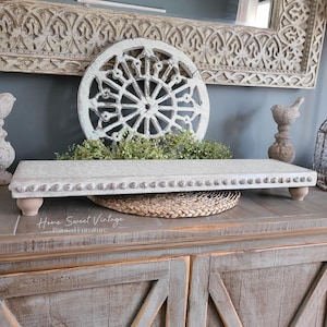 May include: White wooden tray with decorative nail head trim and turned wooden legs. The tray is sitting on a wooden table with a woven straw placemat. The tray is in front of a white and gray carved wooden wall decor.