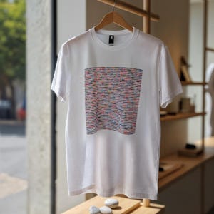 May include: White t-shirt with a colorful rectangular design. The design features horizontal stripes in various colors, including pink, blue, and yellow. The shirt is hanging on a wooden hanger.