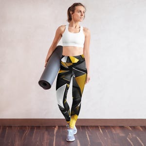 May include: A woman in a white sports bra and patterned leggings. The leggings have a black, white, and gold geometric design. She is holding a rolled-up grey yoga mat. The background is a light beige wall and a wooden floor.