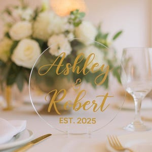 May include: A clear, round acrylic wedding sign with gold lettering that reads "Ashley & Robert EST. 2025." The sign is displayed on a white table, with a floral arrangement in the background. A wine glass and silverware are also visible.
