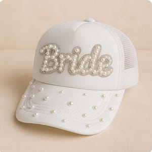 Könnte beinhalten: Weiße Trucker-Cap mit dem Wort "Bride" aus Kunstperlen und Strasssteinen. Der Schirm ist mit zusätzlichen Perlen verziert. Die Rückseite der Kappe ist weißes Mesh.