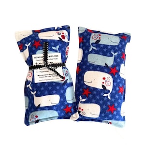 May include: Two blue whale-themed rice-filled pillows. The pillows feature a pattern of white whales, red stars, and blue stars. One pillow is tied with a black ribbon and a tag that reads "HOMEGROWN PILLOWS".