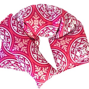 May include: A pink and white patterned neck pillow with a floral design. The pillow is made of fabric and is filled with rice or buckwheat.