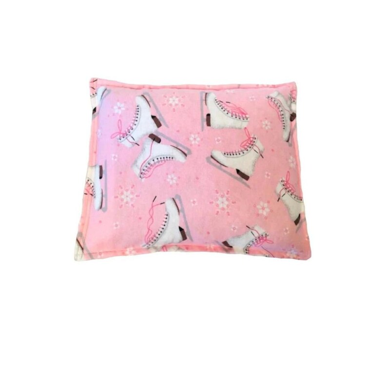 Puede incluir: Una almohada rectangular rosa con un estampado de patines de hielo blancos con cordones rosas y peque&ntilde;os dise&ntilde;os de copos de nieve. La almohada tiene un borde rosa y est&aacute; sobre un fondo blanco.