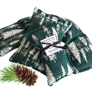 May include: A collection of dark green fabric pillows with a white forest print, tied together with a black ribbon. The pillows are accompanied by a pine cone and a sprig of pine needles. The text on the label reads "Homegrown Pillows".
