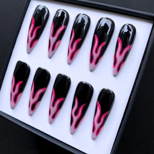 May include: Set of ten long, glossy black press-on nails with a flame design. The flames are painted in shades of pink and light pink, creating a bold and stylish look. The nails are arranged in a black box.