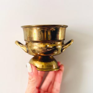 May include: A vintage brass bowl with two small handles and a pedestal base. The bowl has a shiny, aged appearance, suggesting it is an antique or a well-loved item. The design is simple yet elegant, suitable for display or use.