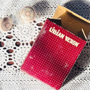 May include: A red box with the text "Lilian Veron" and white polka dots, next to three small, round, beige objects with a circular design. The objects are on a white, crocheted surface.