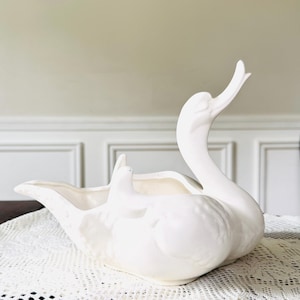 May include: A white ceramic swan-shaped planter with a long neck and open beak. The swan is resting on a white lace doily. The planter has a large opening for plants or flowers.