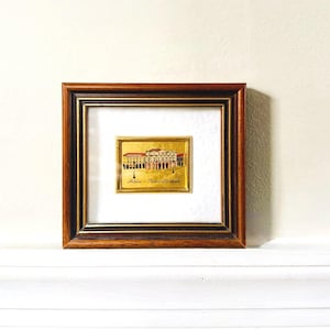 May include: A framed gold foil art piece featuring a building facade with red and white accents. The artwork is set within a dark brown wooden frame with a gold inner border, displayed on a white surface.