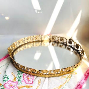 May include: A round vanity tray featuring a reflective mirrored base and an ornate, gold-toned metal gallery rim. The metal edge displays a detailed, openwork floral pattern that encircles the entire perimeter of the circular mirror surface.