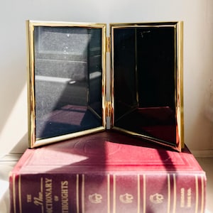 May include: A double-hinged picture frame with a polished gold-tone metal border and two rectangular glass panels. The frame is positioned open in a V-shape, featuring a reflective surface and thin, clean edges surrounding the empty black interior.