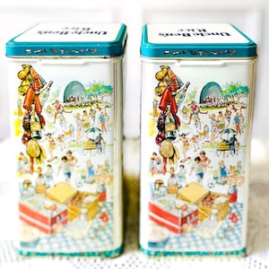 May include: Two vintage Uncle Ben's Rice tins. The tins are white with a teal blue lid and a colorful illustration of a picnic scene. The illustration includes people, food, and bottles. The text "Uncle Ben's Rice" is on the lid.