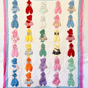 May include: A handmade quilt featuring appliqued figures of children in various outfits. The quilt has a white background with a pink and blue border. The figures are in a variety of colors, including red, blue, yellow, and green. The quilt is likely made of cotton.