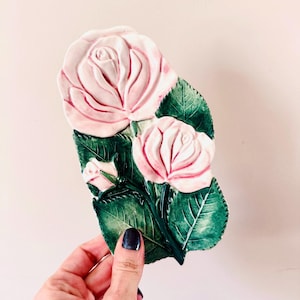 May include: A ceramic rose-shaped decorative piece with three pink roses and green leaves. The roses have detailed petals and are arranged on a green base. The piece is held in a hand.