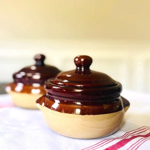 May include: Two small, round ceramic pots with lids, featuring a two-tone design with a glossy dark brown glaze on the upper half and a matte tan finish on the lower half. Each pot has two small side handles and a knob on the center of the lid.