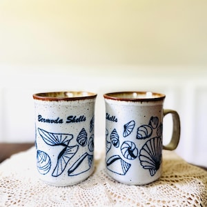 May include: Two ceramic mugs with a speckled off-white glaze and a brown rim. The mugs feature dark blue line drawings of various seashells and the text "Bermuda Shells" printed on the front. Each mug has a sturdy handle and a cylindrical shape.