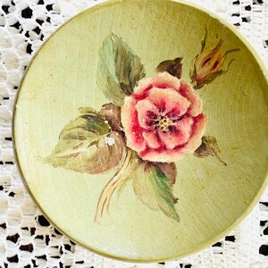 May include: A small, round, antique-style plate with a hand-painted floral design. The plate features a central pink rose with green leaves and a yellow-green background. The plate is likely made of ceramic.