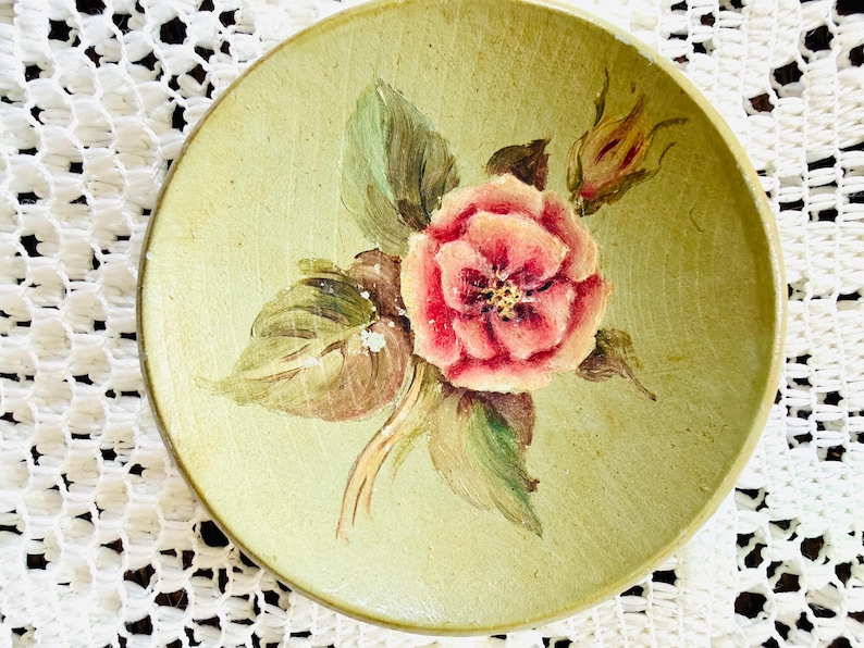May include: A small, round, antique-style plate with a hand-painted floral design. The plate features a central pink rose with green leaves and a yellow-green background. The plate is likely made of ceramic.