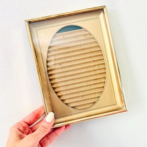 May include: A gold-colored picture frame with an oval opening. The frame has a glass front and a corrugated cardboard backing. The frame is held by a hand against a white background.